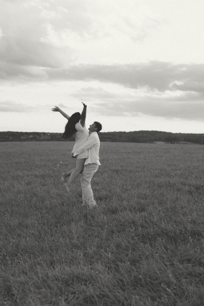 Couple embracing and lifting each other in a grassy field beneath a cloudy sky in a black and white engagement photo.