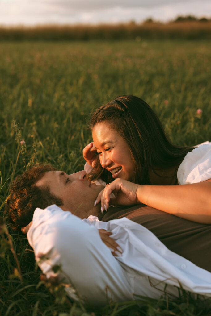 Close up of a couple lying in a grassy field, laughing together in warm golden hour light.