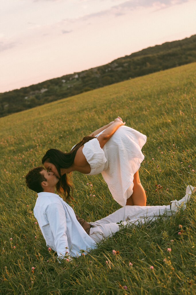 Couple kissing in a grassy field as the woman bends down toward her partner, surrounded by warm evening light.
