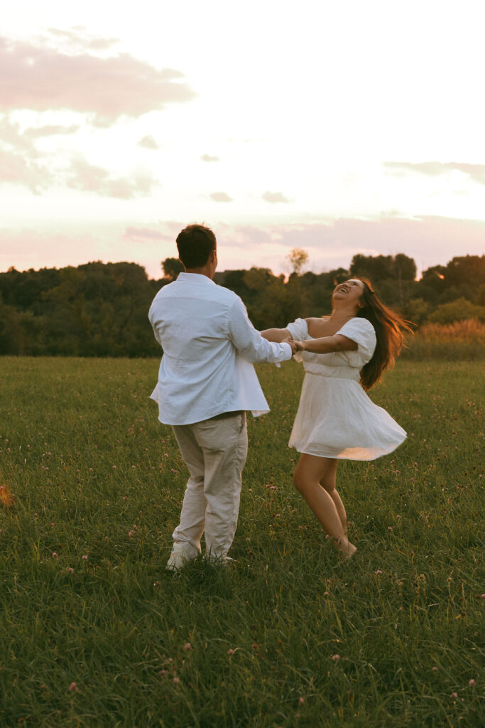 Couple holding hands and dancing playfully in a grassy field at sunset, with the woman laughing in her flowing white dress.