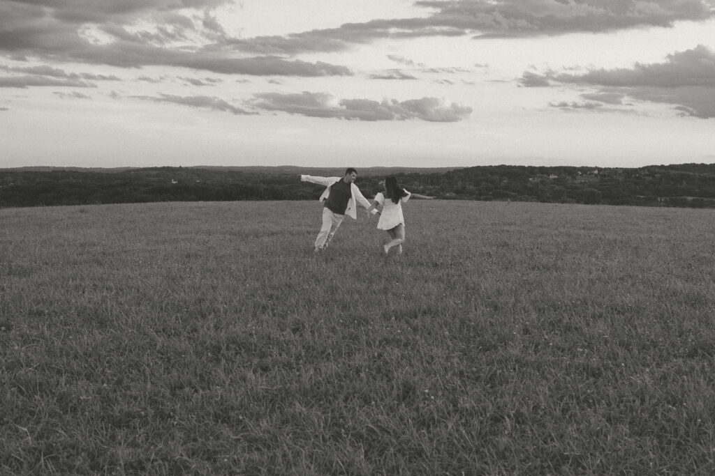 Couple holding hands and running through a grassy field beneath a cloudy sky in black and white.