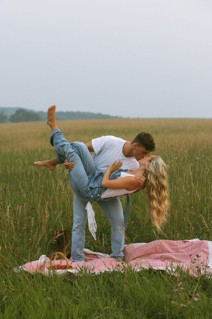 Couple sharing a kiss beside a red gingham picnic blanket in a Wisconsin field